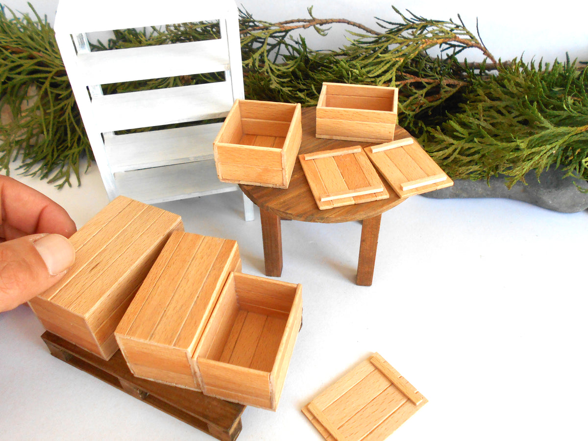 A hand is holding a miniature wooden crate in the foreground, with several small wooden storage boxes and lids arranged on a round table. The scene includes a white shelving unit and green foliage in the background, suggesting a dollhouse or scale model furniture setup.