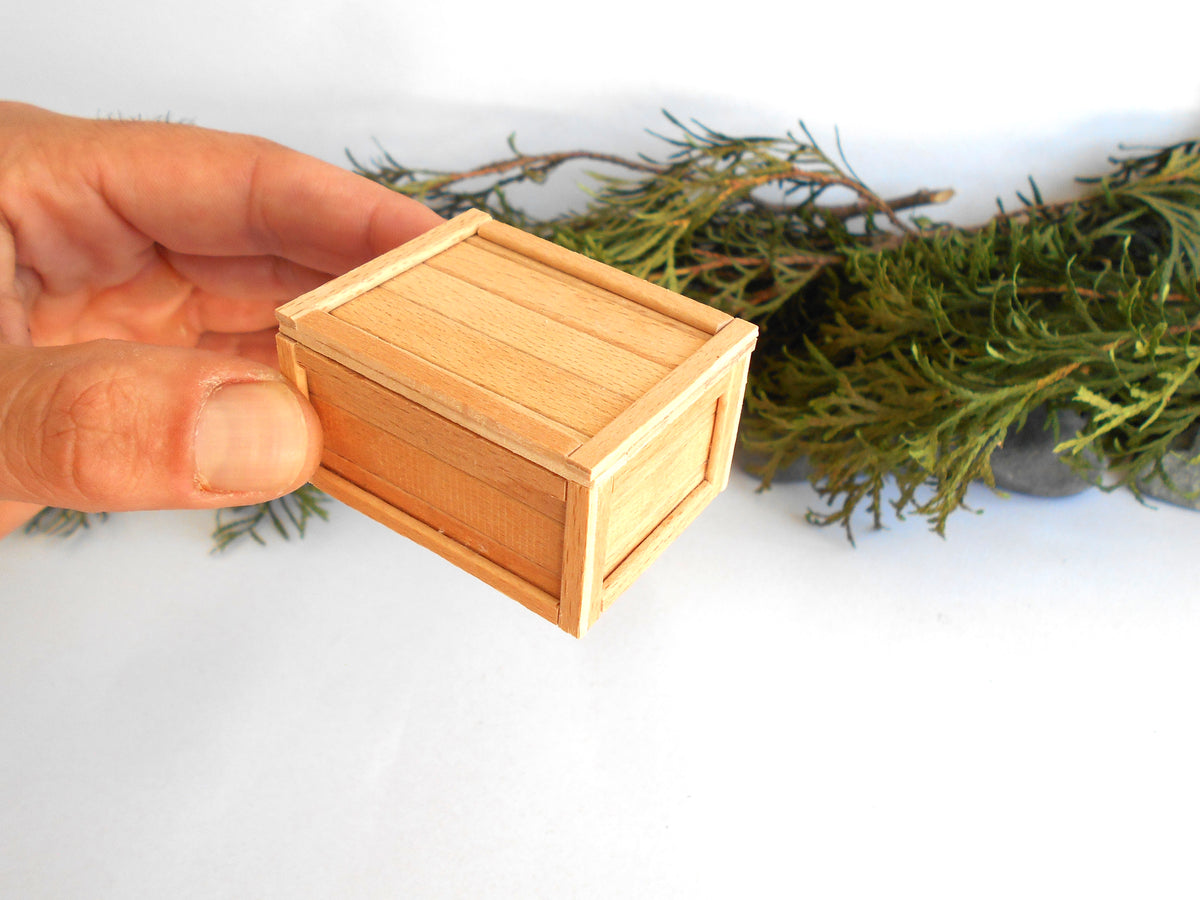 A hand holding a small, light-colored wooden box with a visible wood grain, set against a white background with green pine branches in the background.