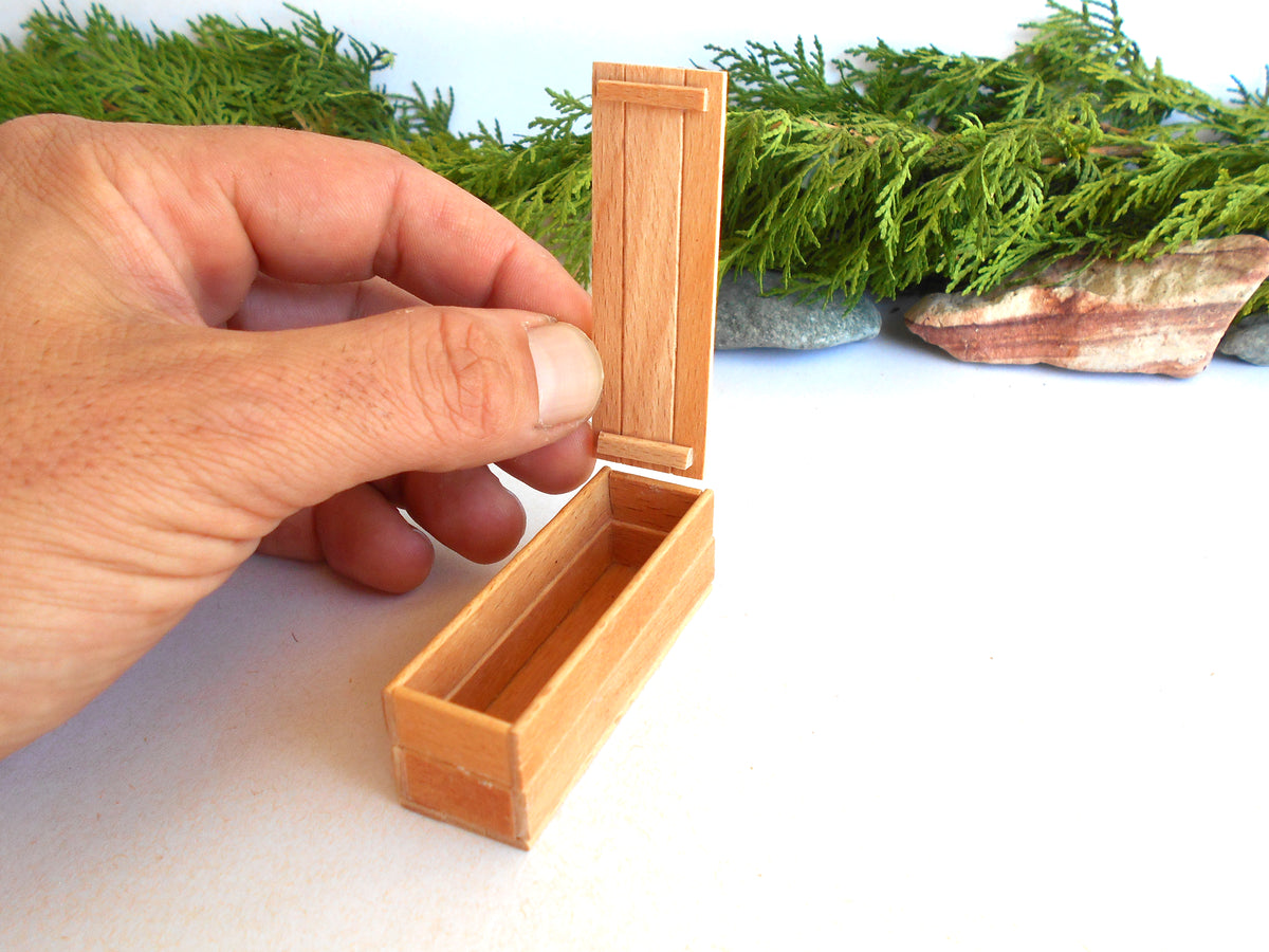 A hand holding open a small wooden box with a hinged lid, set against a white background with green foliage and stones in the blurred background.