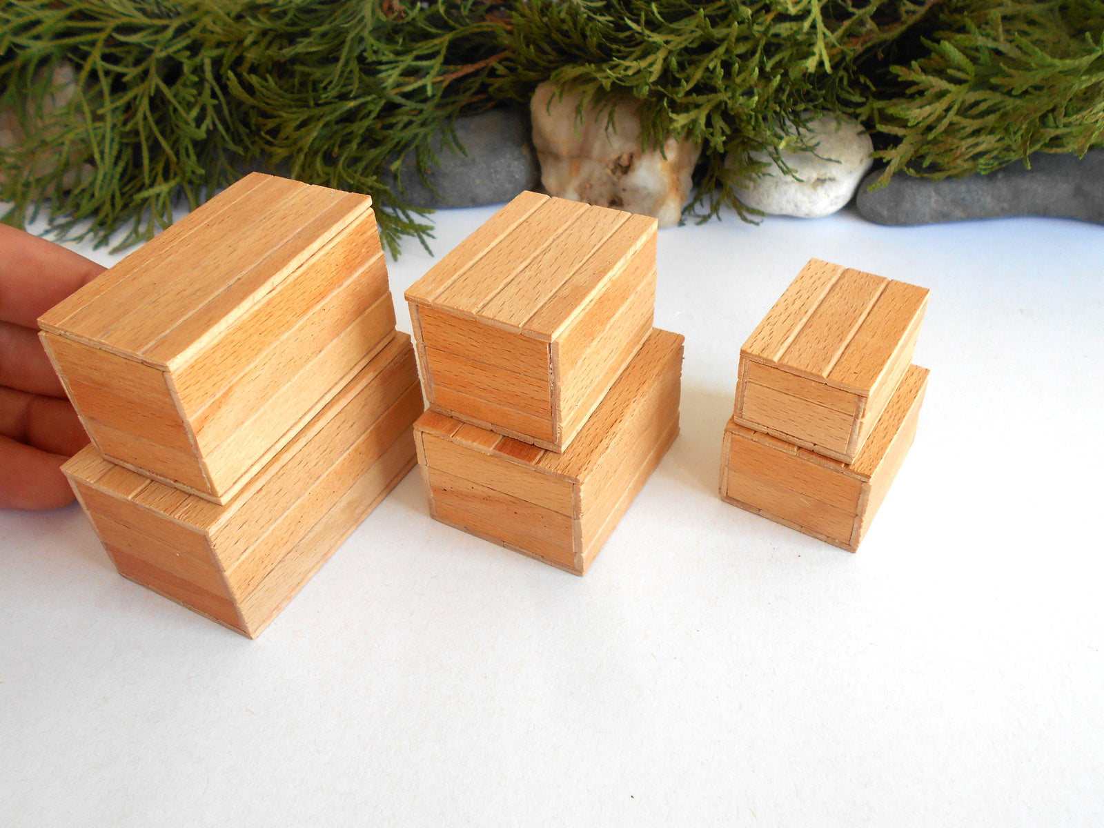 Four small rectangular wooden miniature boxes in different sizes, stacked on a white surface with green foliage and stones in the background. A hand is visible on the left holding one of the larger miniature boxes .