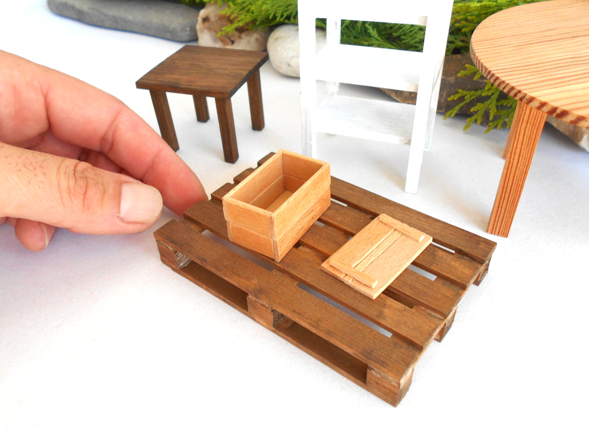 A miniature wooden crate set with a lid, displayed on a small wooden pallet base. A human hand is visible on the left holding the pallet, and other miniature furniture pieces are blurred in the background.