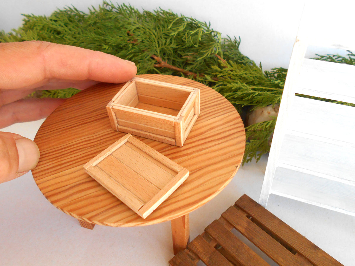 A close-up of a miniature wooden box set on a small round wooden table, with a hand holding one of the boxes and green foliage in the background.