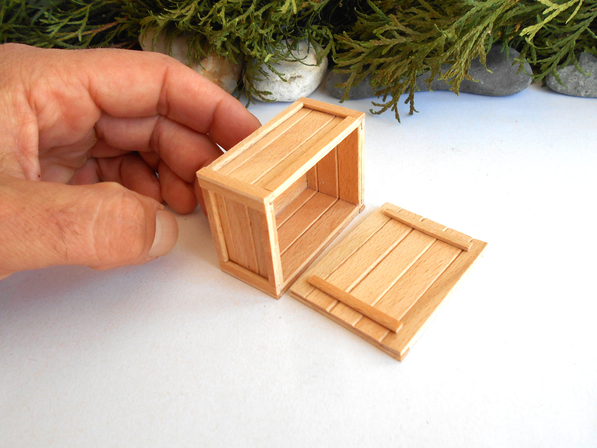 A miniature wooden crate model with its lid off, held by a person's hand on a white surface with decorative moss and stones in the background.