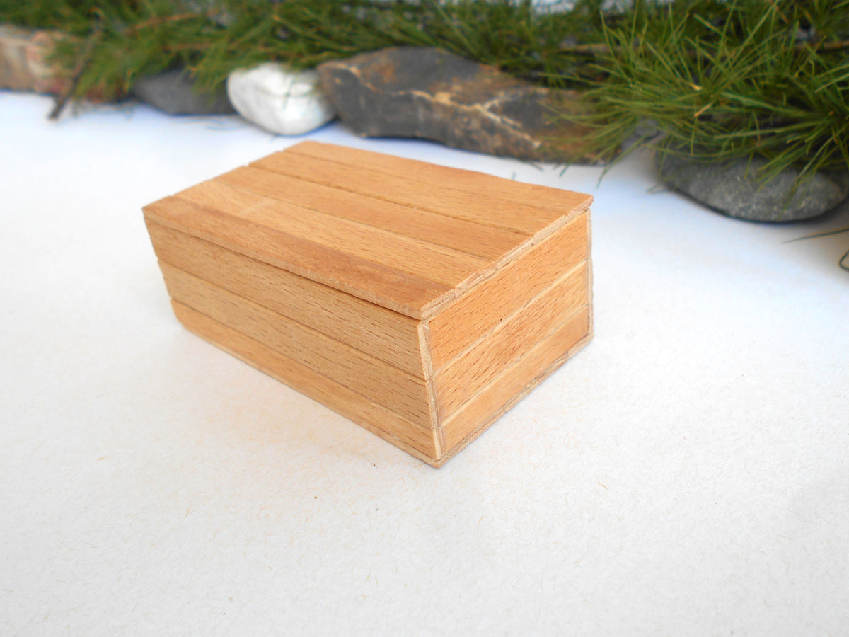 A small rectangular wooden box with a hinged lid, made from light natural wood, shown on a white surface with a blurred background of greenery and rocks.