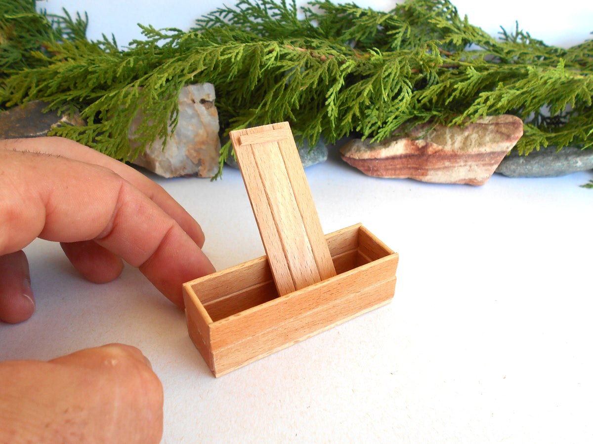 A miniature light wood box with a hinged lid, shown with a hand pointing at it against a white background with green foliage and stones in the blurred background.