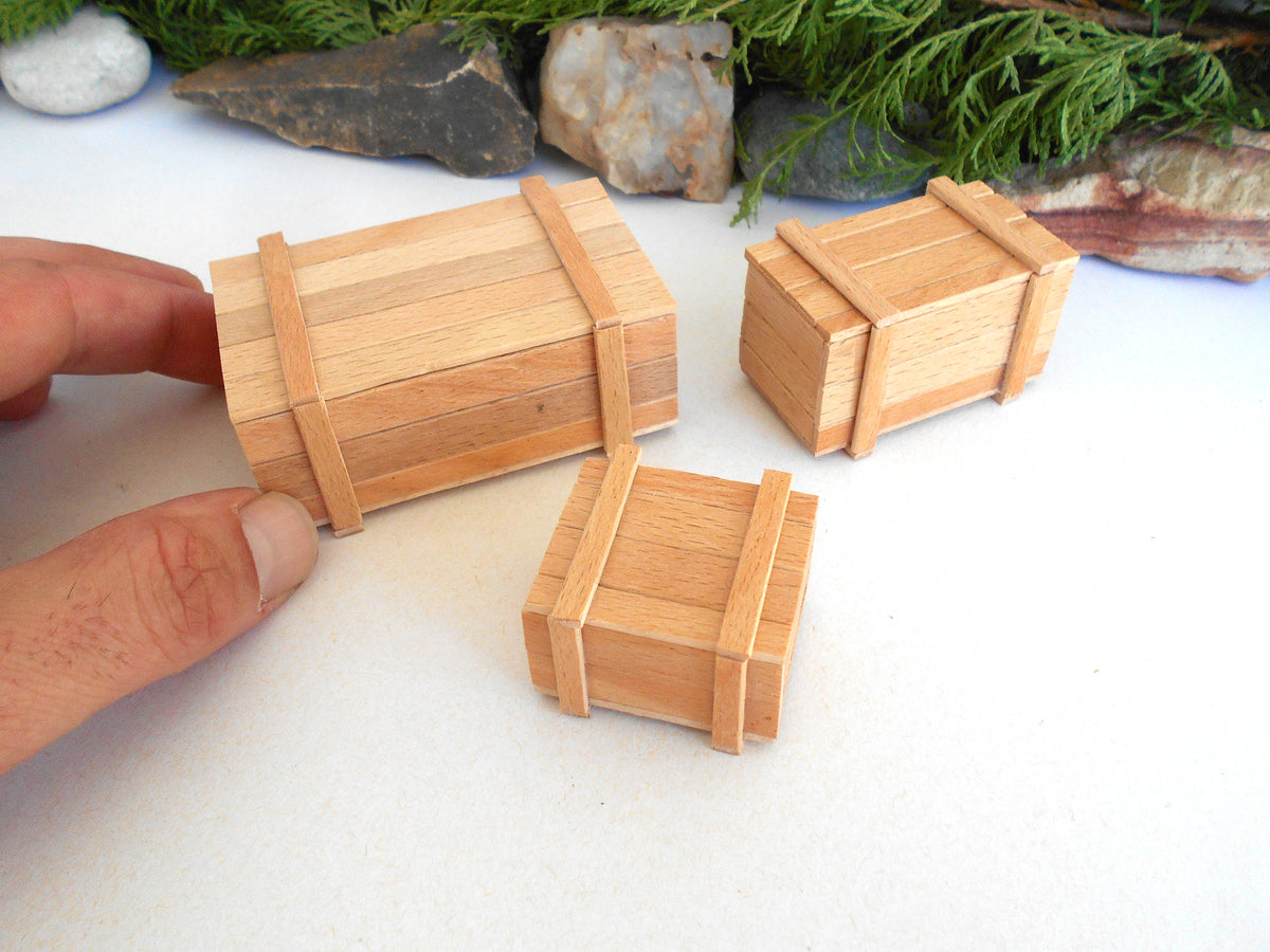 Three miniature wooden crates of different sizes with light brown wood grain and darker brown straps, shown on a white surface with a hand holding one of the larger crates.
