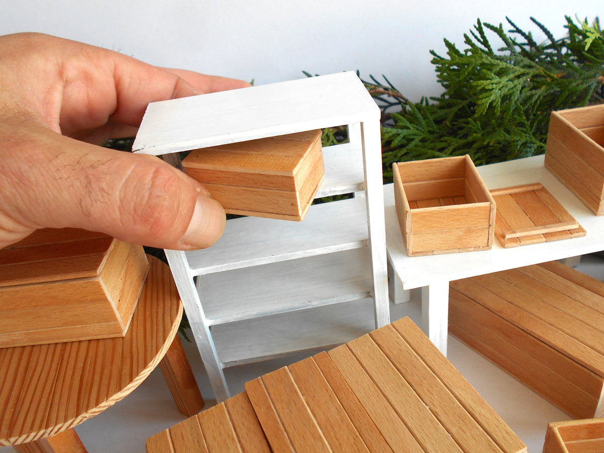 A close-up of a hand holding a small wooden drawer, surrounded by matching miniature light wood furniture pieces and shelves on a white tabletop.