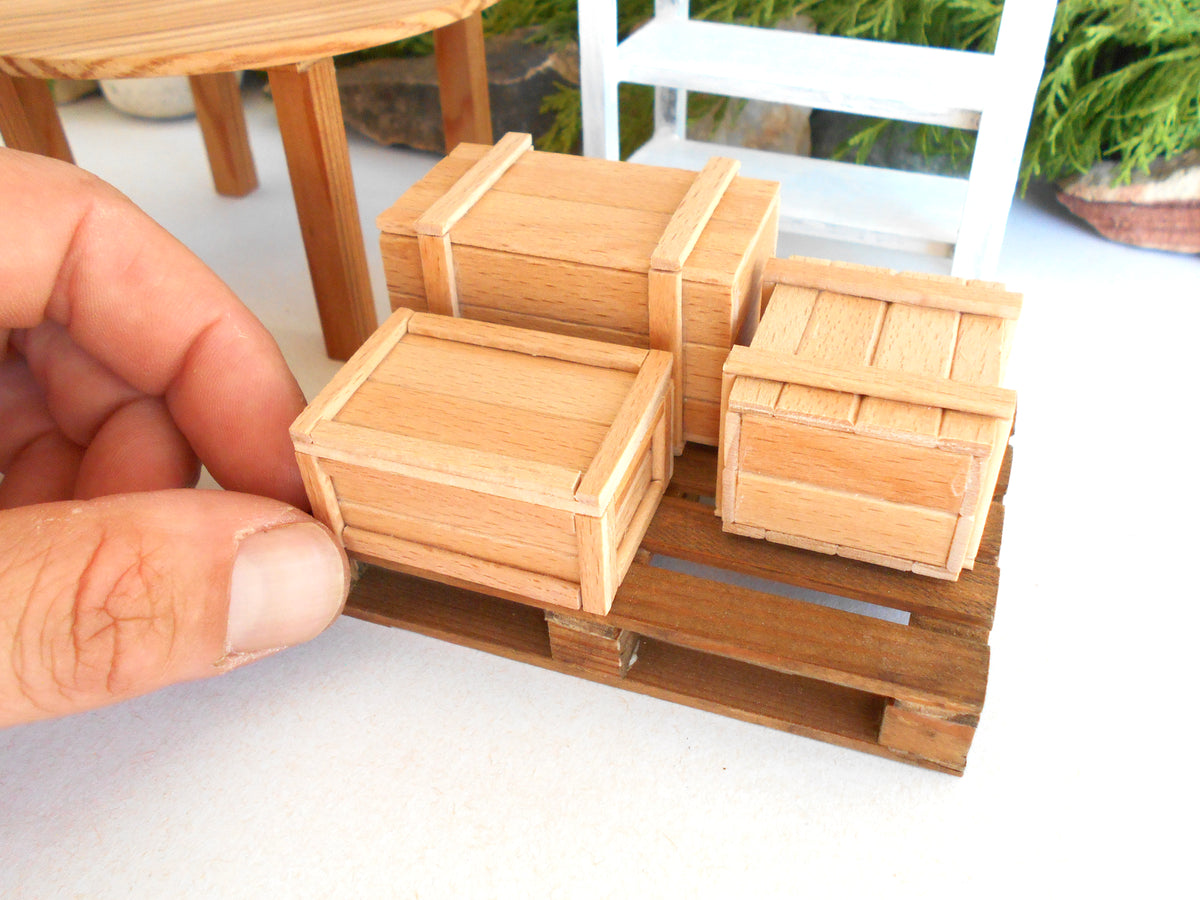Three miniature wooden crates on a small wooden pallet, with a human hand adjusting the front crate in a miniature outdoor-style scene.