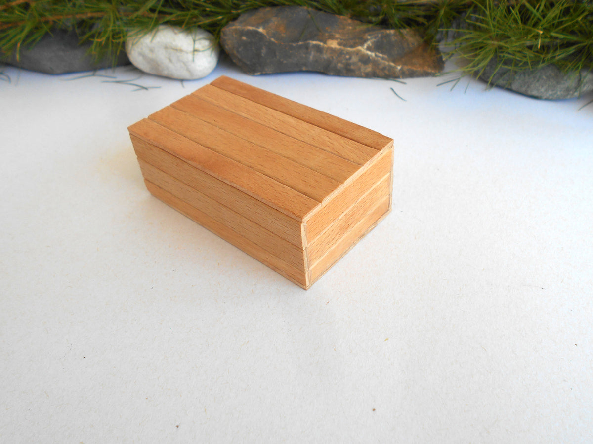A small rectangular wooden box with a slatted lid, shown on a white surface with decorative stones and greenery in the background.