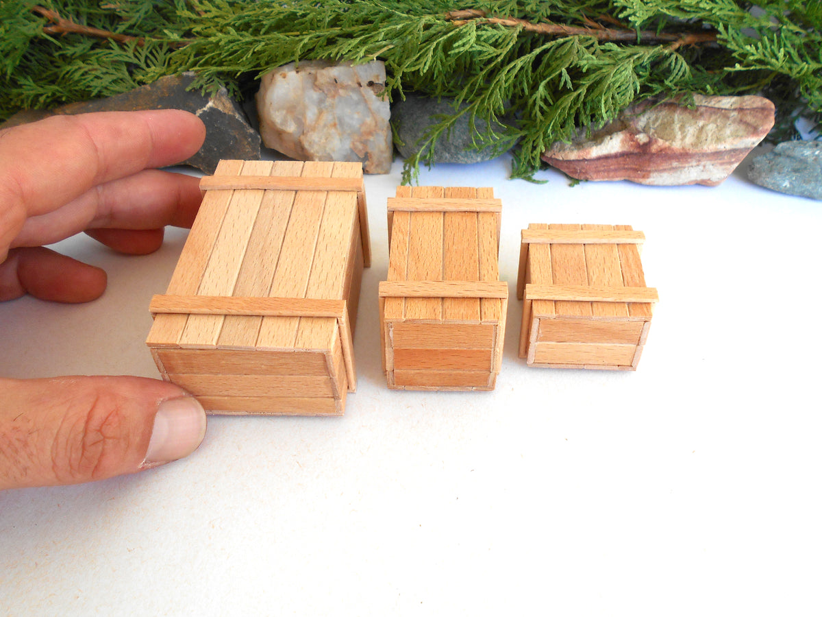 Three miniature wooden crates in different sizes displayed on a white surface, with a hand holding the largest crate for scale and green foliage and rocks in the background.