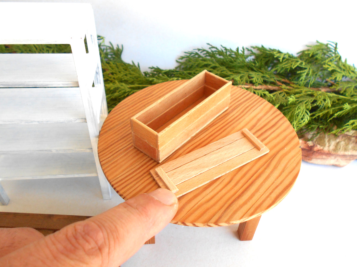 A miniature wooden box set with a lid, displayed on a small round wooden table. A human finger is pointing at the product, indicating its small scale, with a white shelf and green foliage in the background.