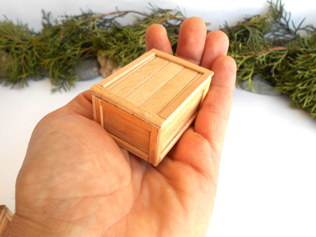 A miniature light wood crate box held in a person's hand, with green foliage blurred in the background.