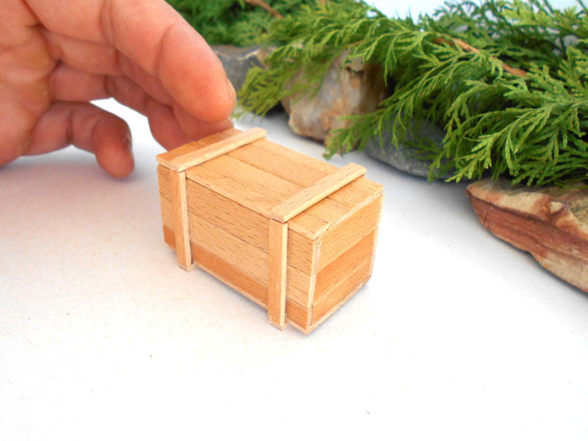 A miniature light wood crate or box with wooden bands, held by a person's hand against a white background with green foliage and rocks in the blurred background.