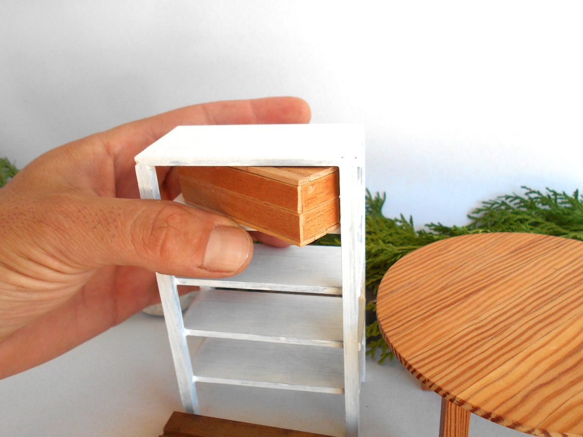 A miniature white shelving unit with a wooden drawer front, held by a hand to show scale next to a round wooden table.