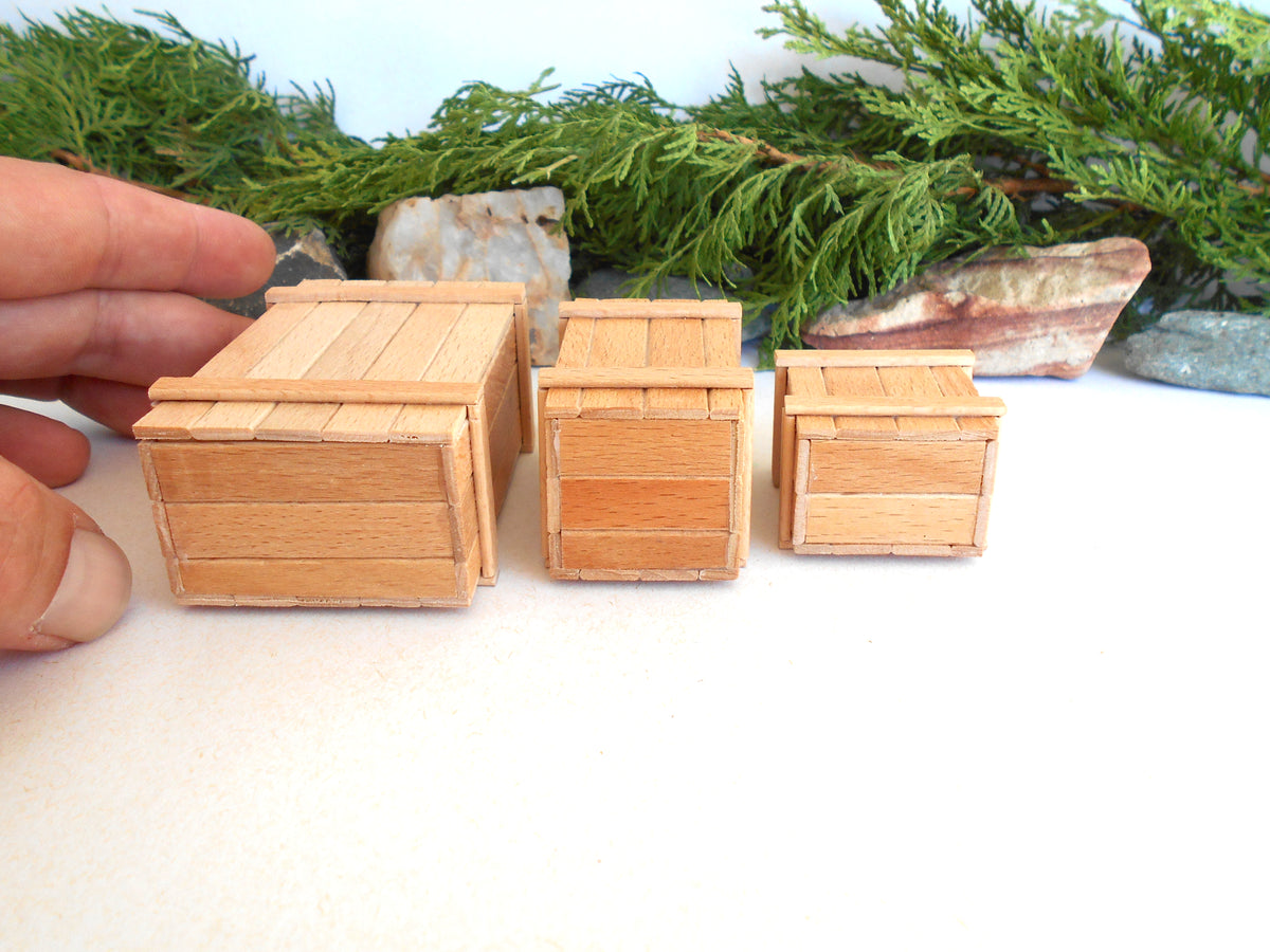 Three miniature wooden crates in different sizes arranged on a white surface, with a hand holding the leftmost crate for scale and green foliage in the background.