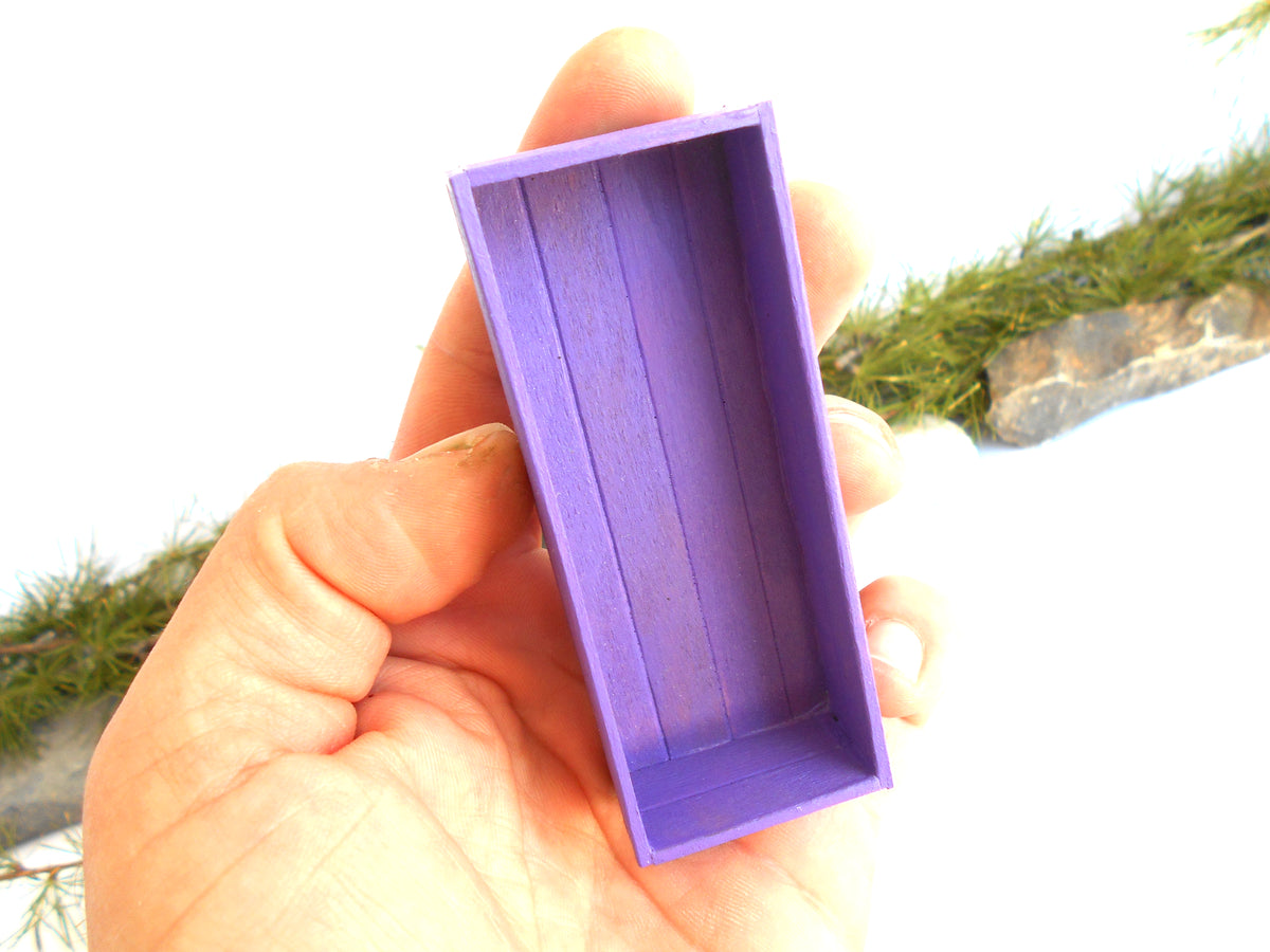 A hand holding a small, empty rectangular purple box with a slanted lid, set against a bright white background with some blurred greenery.