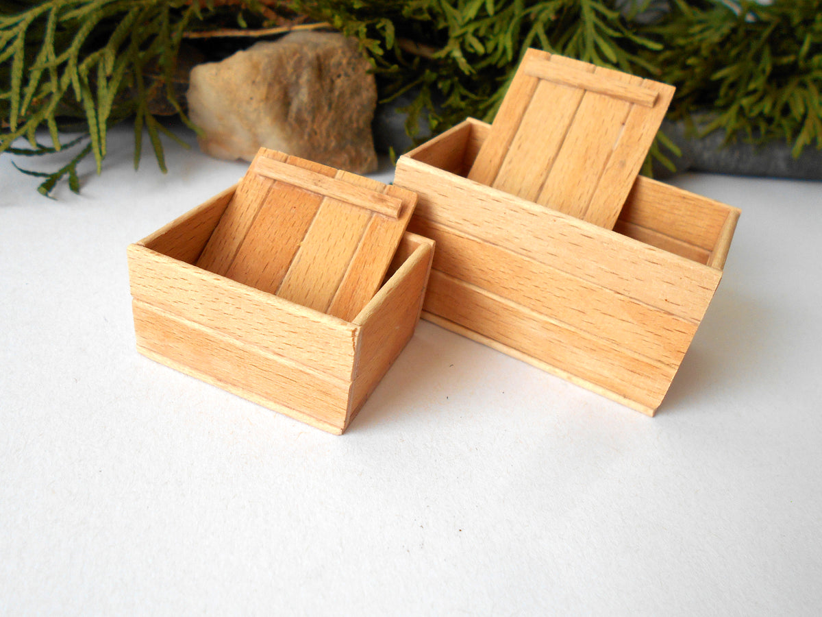 Three miniature wooden boxes with slatted fronts and hinged lids, arranged on a white surface with green foliage and a small stone in the background.