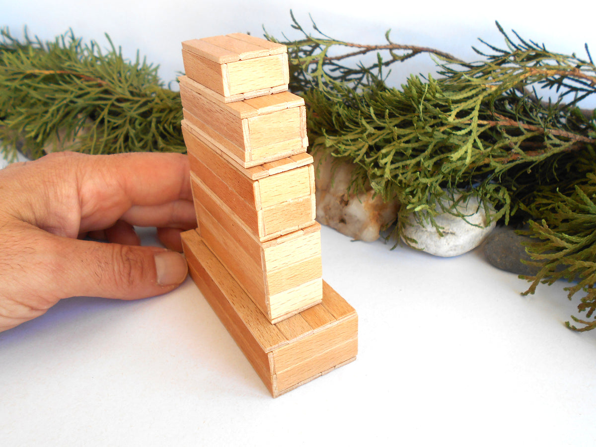 A hand holds a small wooden miniature boxes tower toy made of stacked rectangular wood pieces on a white surface, with green foliage and stones in the background.