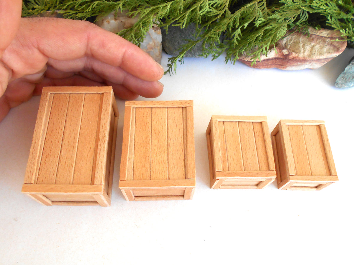 Four miniature light wood crate models in different sizes arranged on a white surface, with a hand holding the largest crate for scale and green foliage in the background.