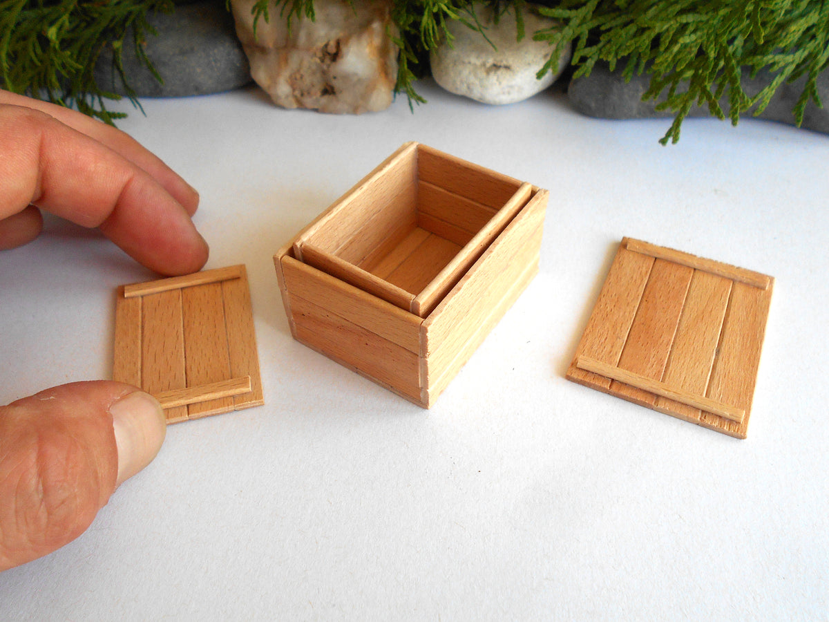 A miniature wooden box with a sliding lid, shown with a hand holding the lid. The box is made of light-colored wood with a slatted front and side panels, set against a white background with decorative greenery and stones.