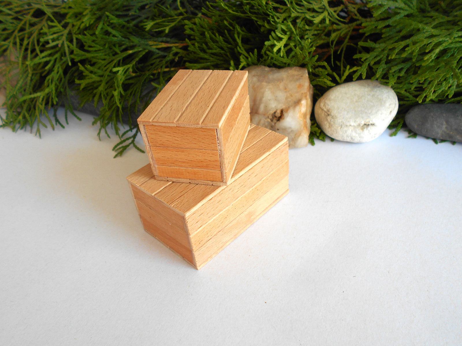 Two small stacked wooden boxes with a natural light brown finish, styled on a white surface with green foliage and smooth stones in the background.