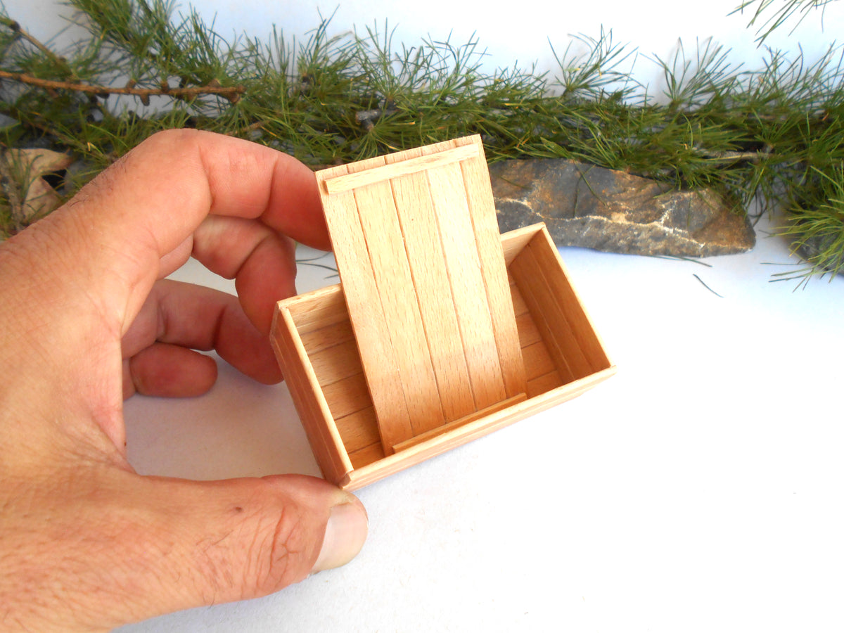 A miniature light wood box with a hinged lid held open by a person's hand, set against a white background with pine branches and a small stone prop.