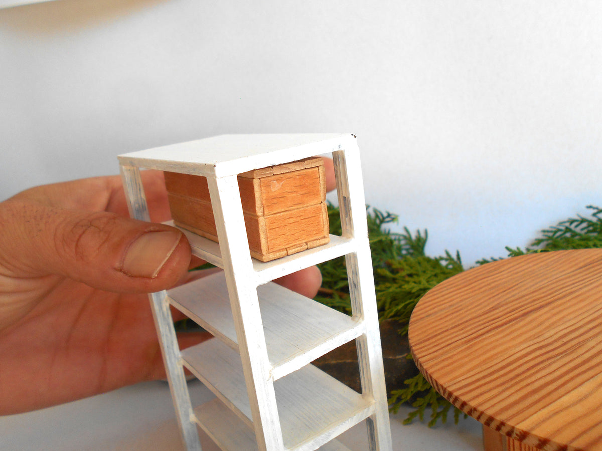 A hand holding a miniature white shelving unit with a small wooden chest on the top shelf, shown in a close-up product photo.