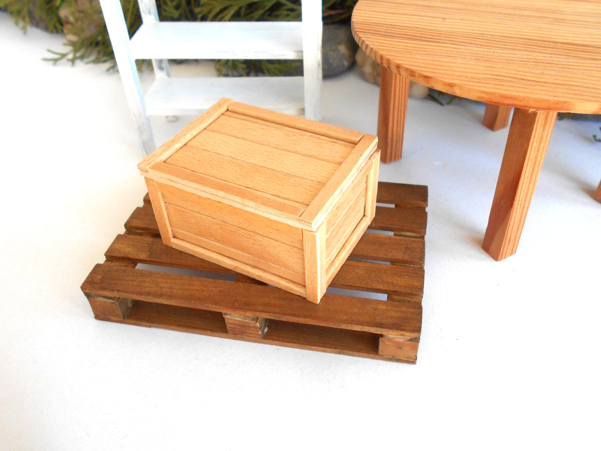A small wooden storage box with a slatted lid sits on a miniature brown wooden pallet, set against a white background with miniature furniture props.