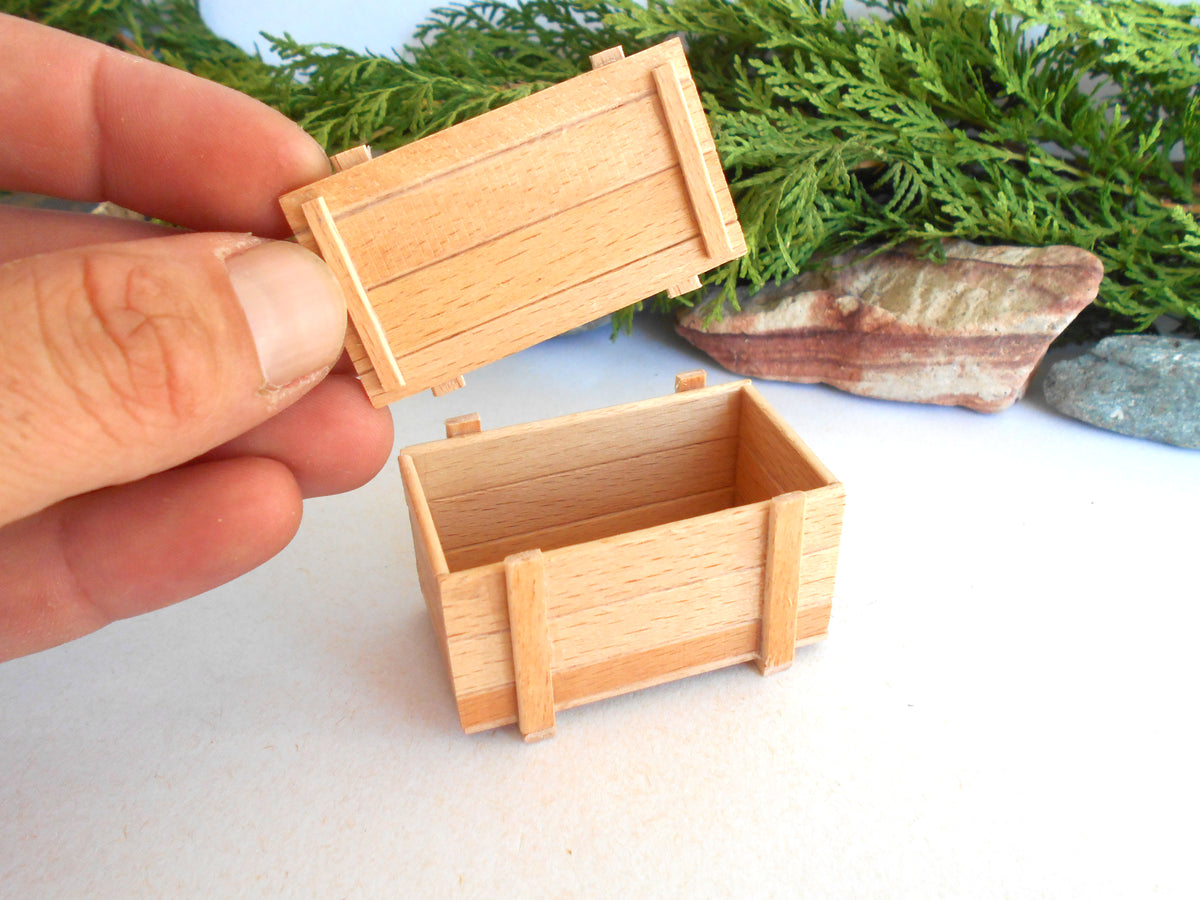 A hand holding the lid of a small unfinished wooden box next to the open box base, with green foliage and stones in the background.