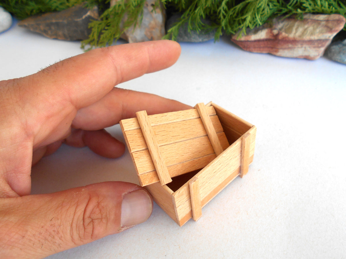 A miniature wooden crate or box being held by a person's hand, shown close-up on a white surface with blurred rocks and greenery in the background.