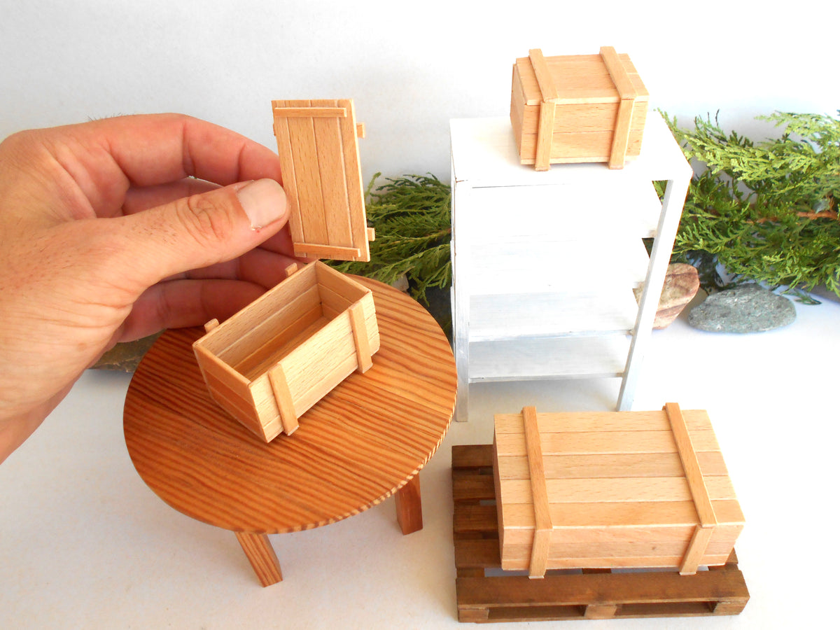 A hand holds a miniature wooden crate door next to a small wooden table with a miniature wooden crate on top, with other miniature wooden storage boxes and a white shelf in the background.