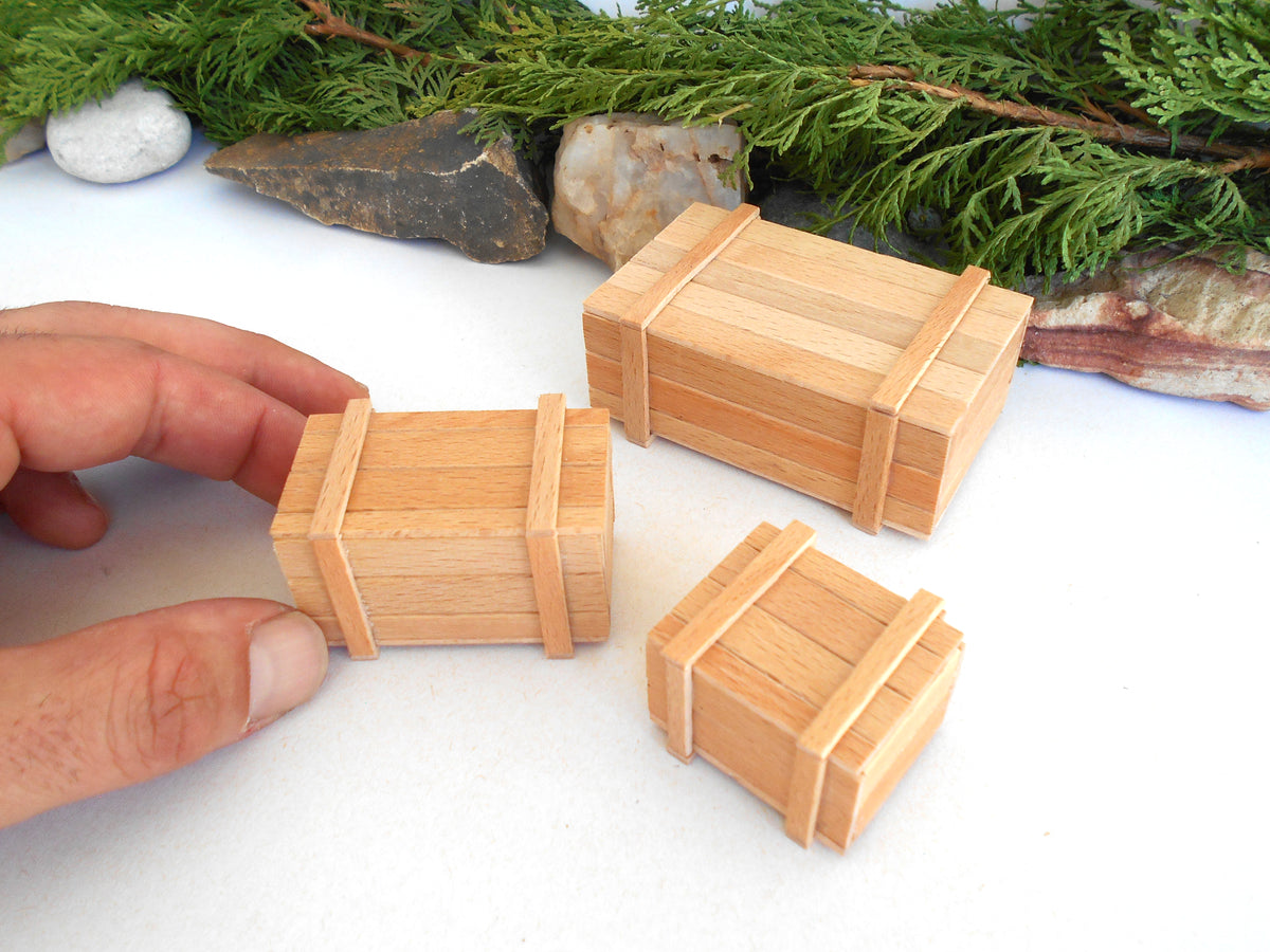 Three miniature wooden crates of different sizes displayed on a white surface with green foliage and rocks in the background. A hand is holding the smallest crate on the left.