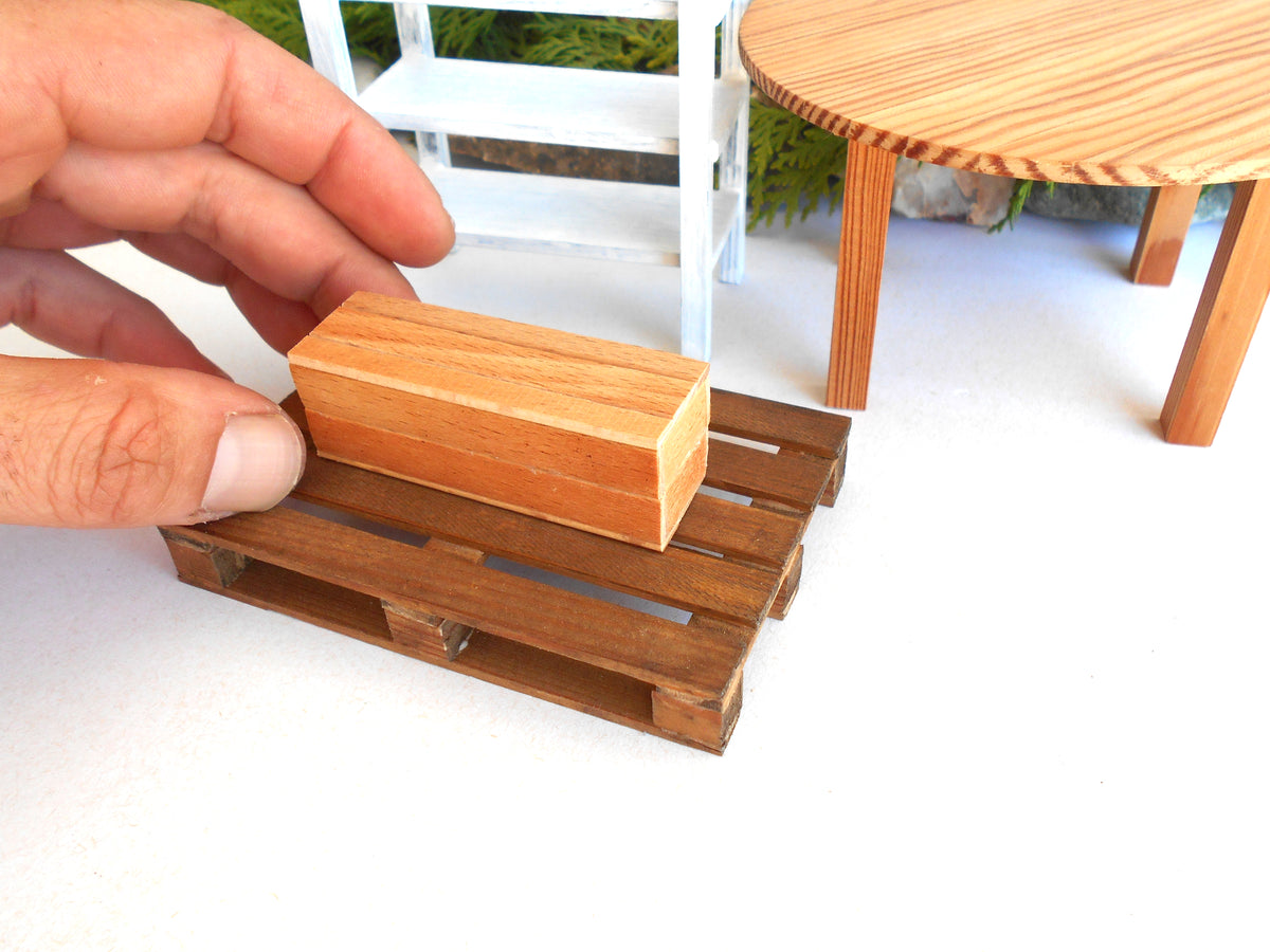 A hand holding a small rectangular wooden miniature box above a miniature brown wooden pallet, with a miniature white chair and round wooden table in the background.