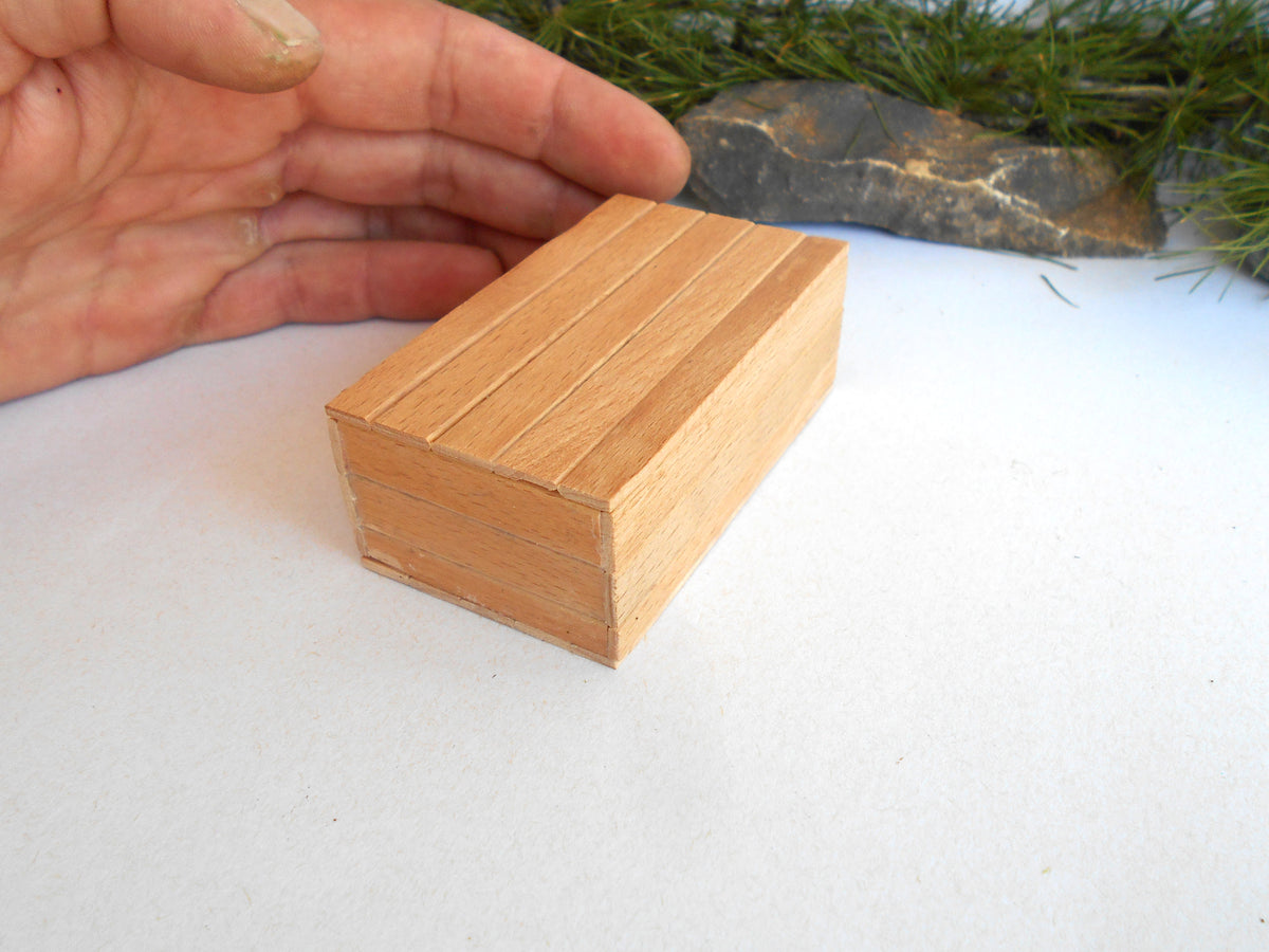 A miniature wooden box with a lid, detailed with vertical grooves, shown next to a person's hand for scale on a white surface with a decorative rock in the background.