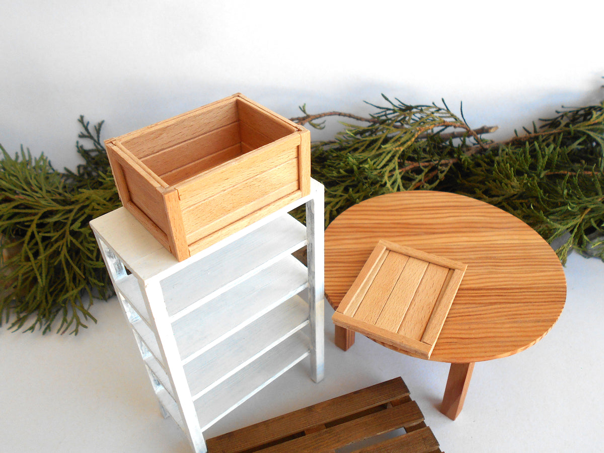 A miniature dollhouse furniture set featuring a round wooden table, a white shelving unit, and a wooden crate, styled with green pine branches on a white background.