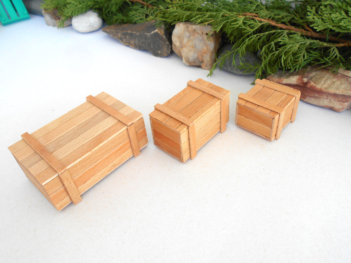 Three miniature wooden crates of different sizes arranged diagonally on a white surface with decorative greenery and stones in the background.