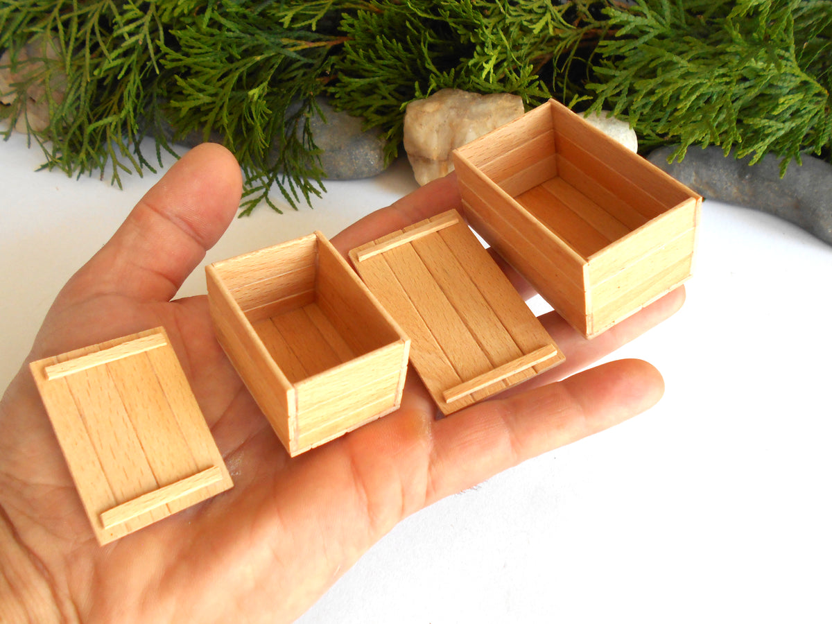 Four miniature light wood boxes of varying sizes held in a person's hand, with green foliage and small stones in the background.