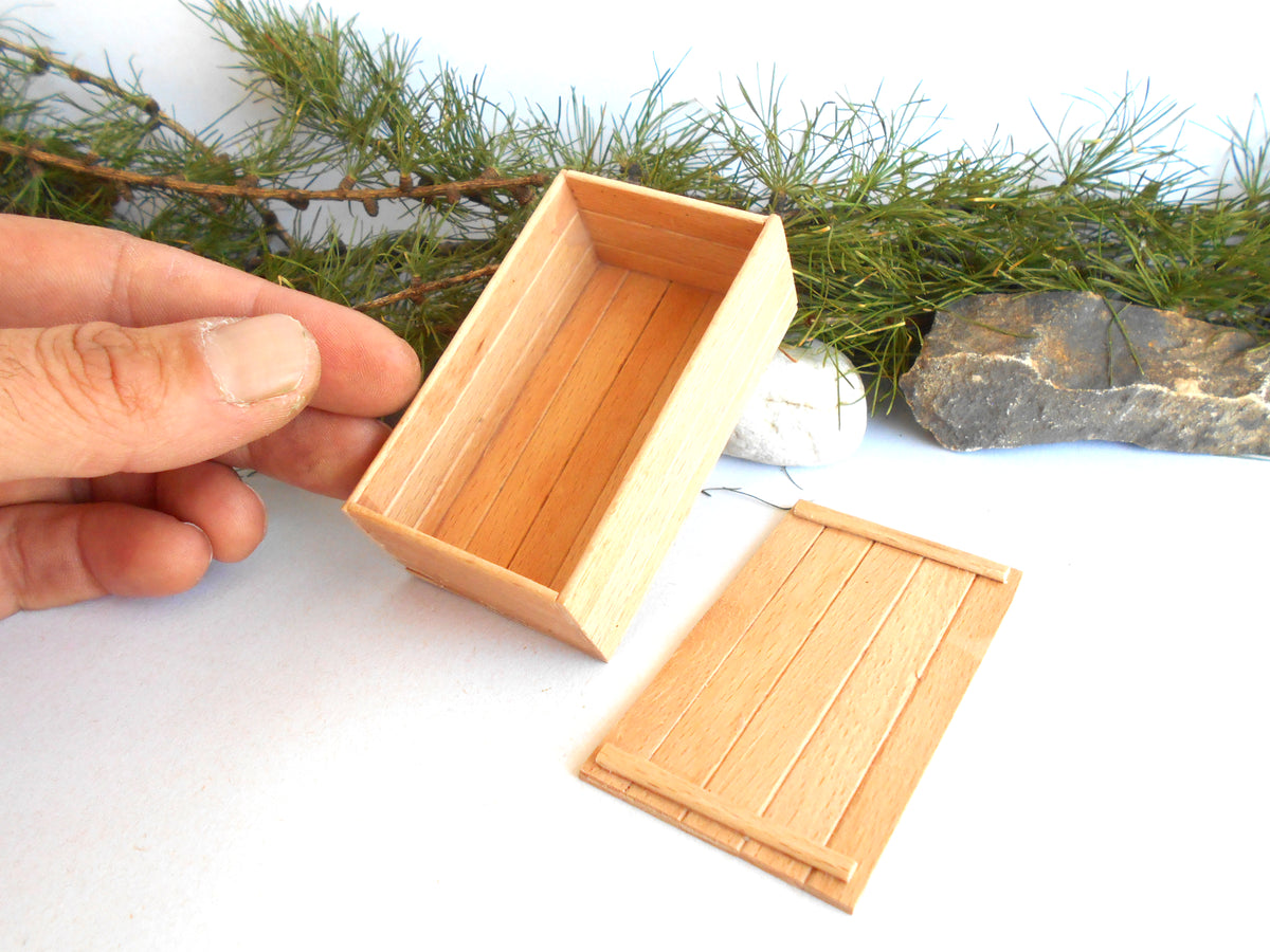 A miniature wooden box with a matching lid, shown in a scale model context with a hand holding the open box. The background features decorative pine branches and a rock on a white surface.