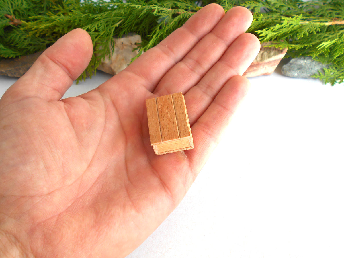 A miniature wooden book-shaped object resting on an open hand, with green foliage and stones blurred in the background.