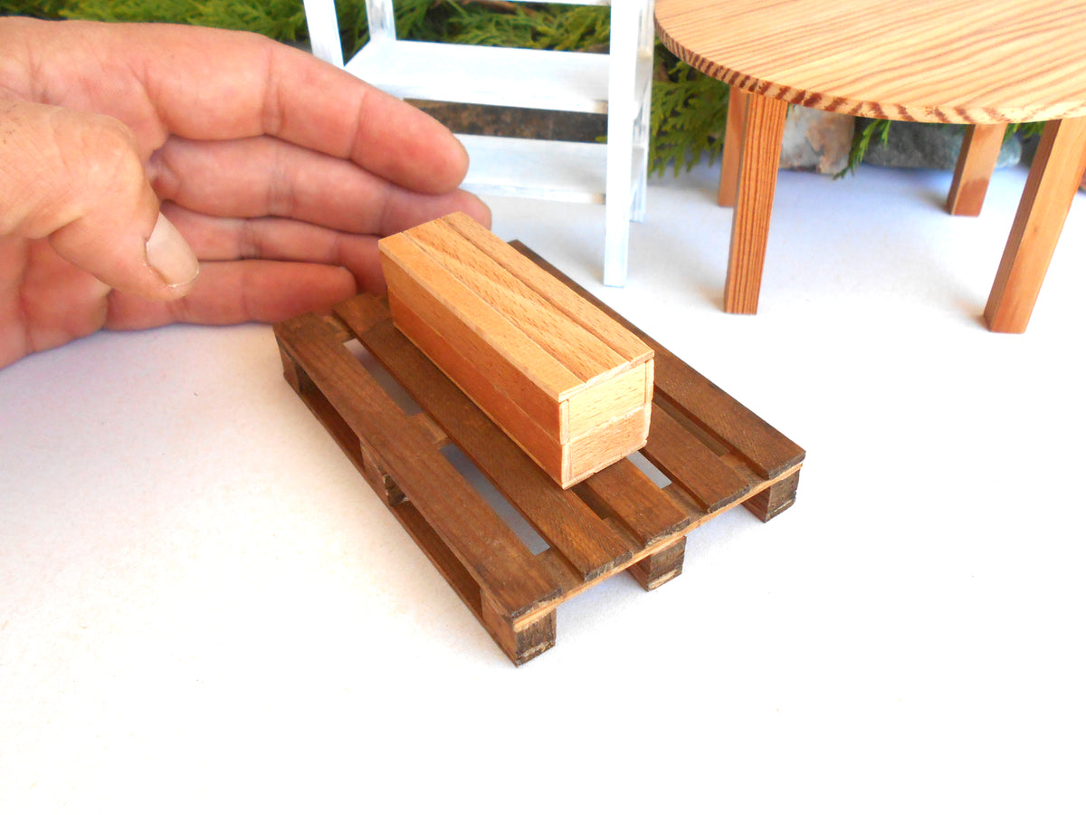 A miniature wooden pallet and matching rectangular miniature box, likely a scale model or dollhouse furniture piece, shown with a human hand holding it for scale against a white tabletop with miniature furniture in the background.