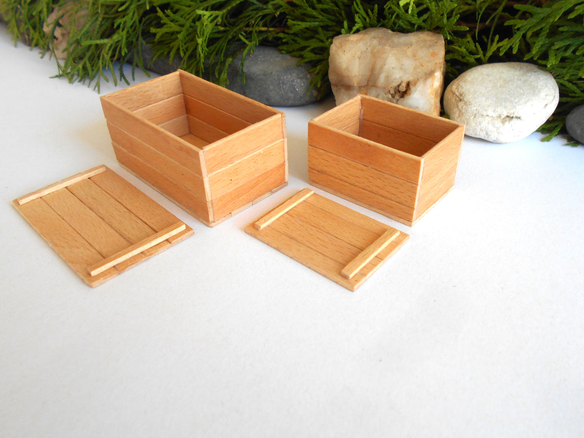 Two small light wood boxes with matching lids, arranged on a white surface with greenery and stones in the background.