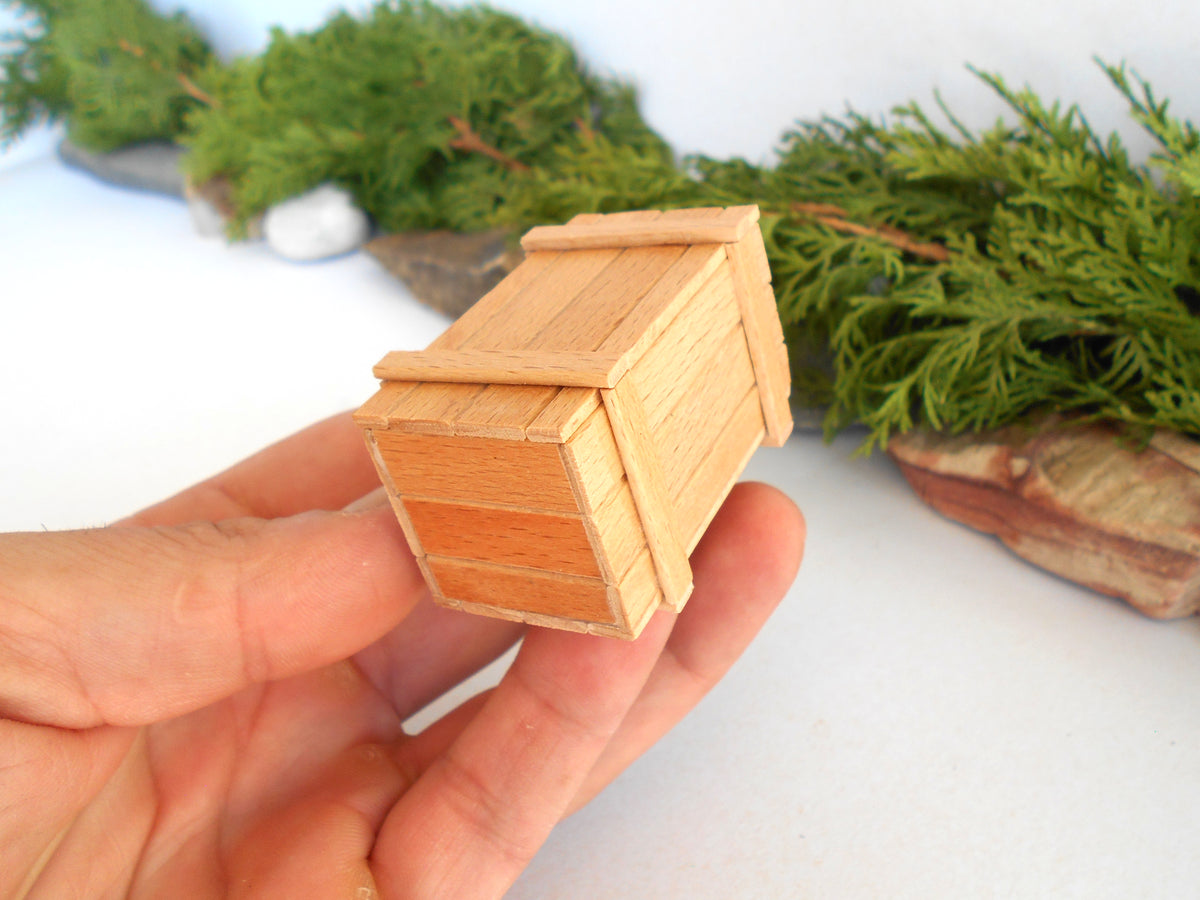 A miniature wooden box, likely a dollhouse or scale model, held in a person's hand against a white background with blurred green foliage.