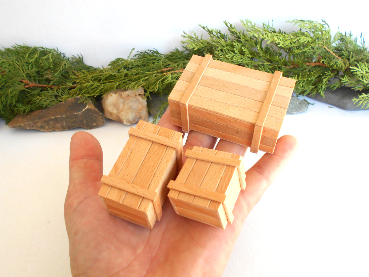 Three miniature wooden crates with metal-looking band straps, held in a person's hand against a white background with greenery and rocks behind them.