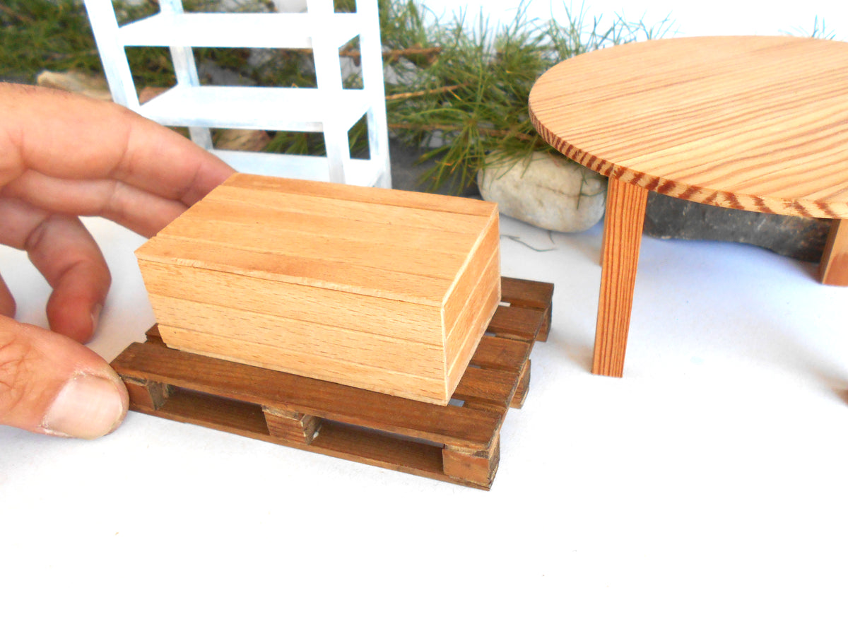 A miniature wooden box with a light natural wood finish sits on a small dark brown wooden pallet, held by a person's hand. The miniature setup is on a white surface with other miniature furniture pieces, including a round wooden table and a white shelf, blurred in the background.