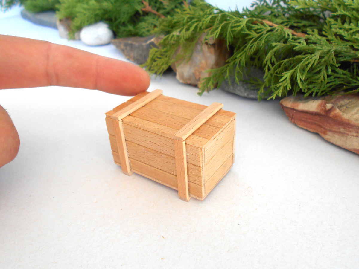 A miniature wooden crate toy or model shown on a white surface, with a human finger pointing at it for scale and green foliage in the blurred background.
