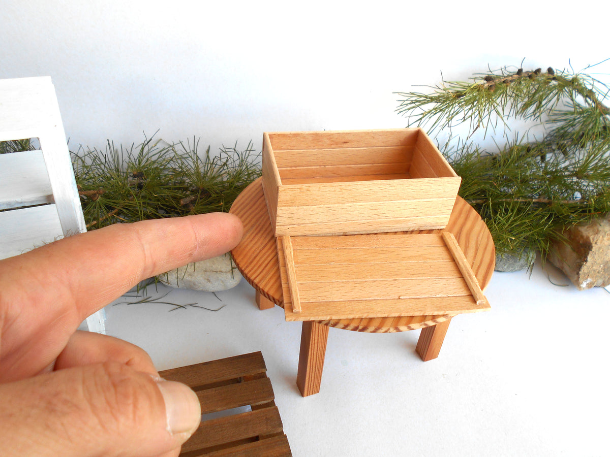A miniature wooden box and tray set displayed on a small round table, with a human hand pointing toward it for scale against a white background with pine branches.