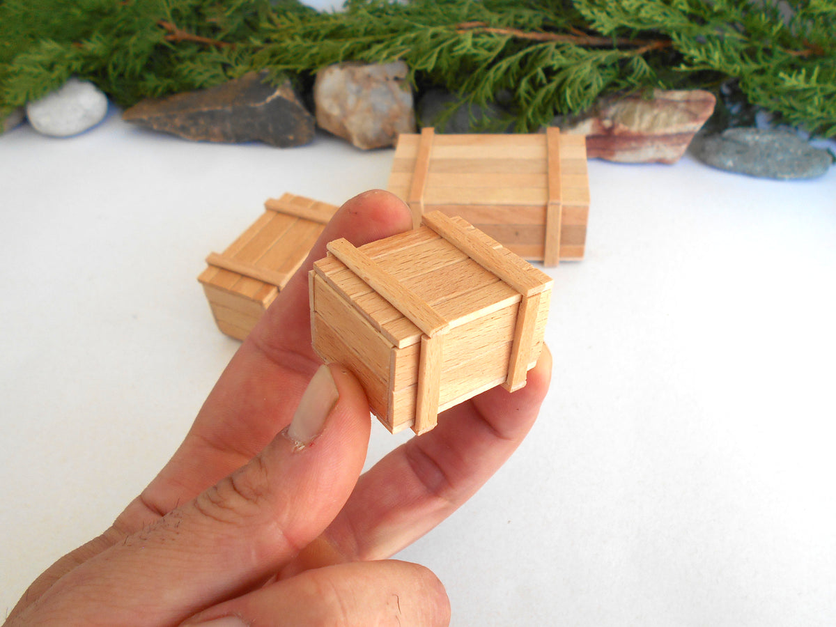 A hand holding a miniature wooden crate box, with two more similar boxes in the background on a white surface with decorative greenery and stones.