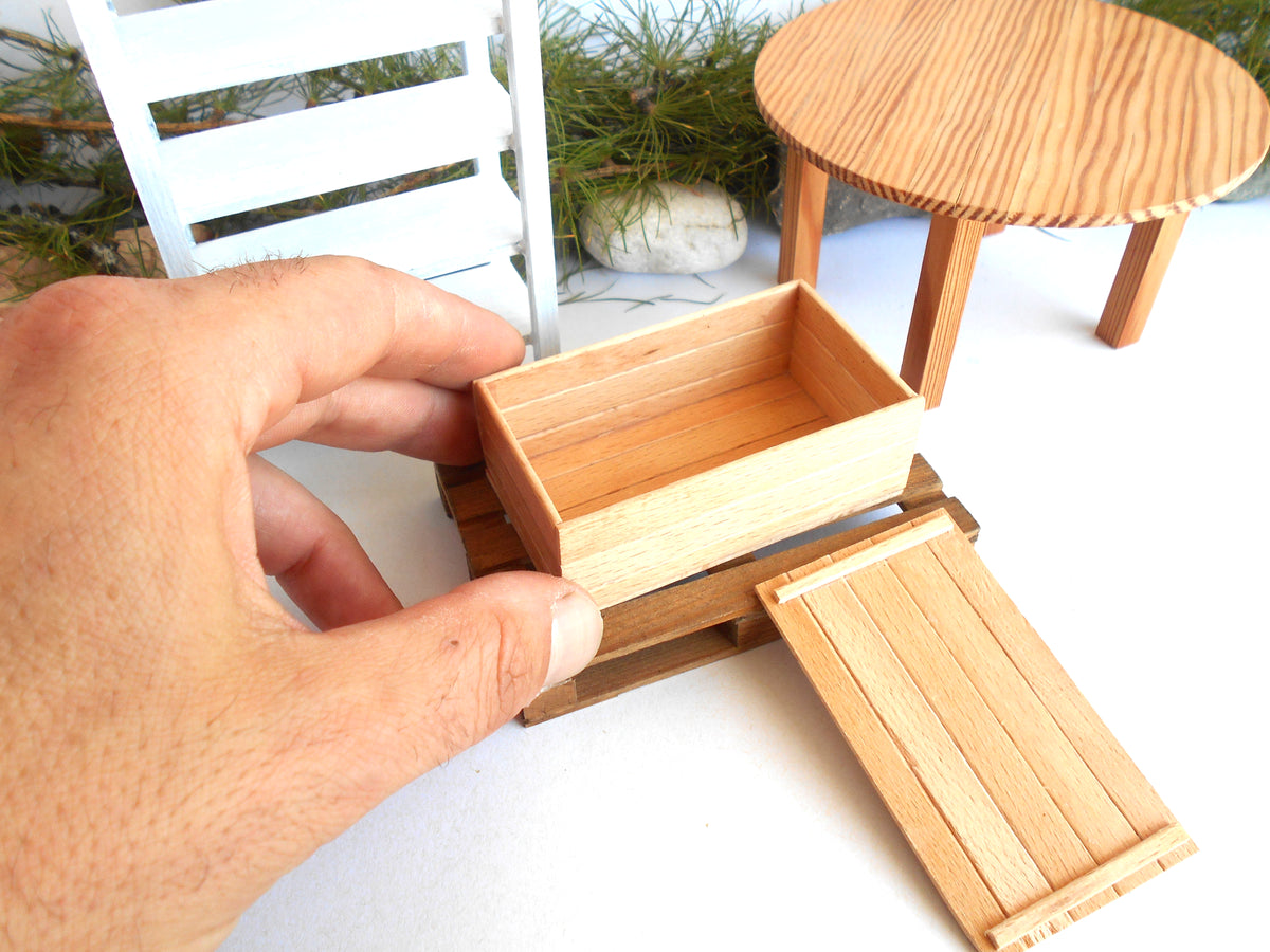 A hand holds a miniature light wood box in front of a dollhouse-style outdoor scene with a small round table and chair.