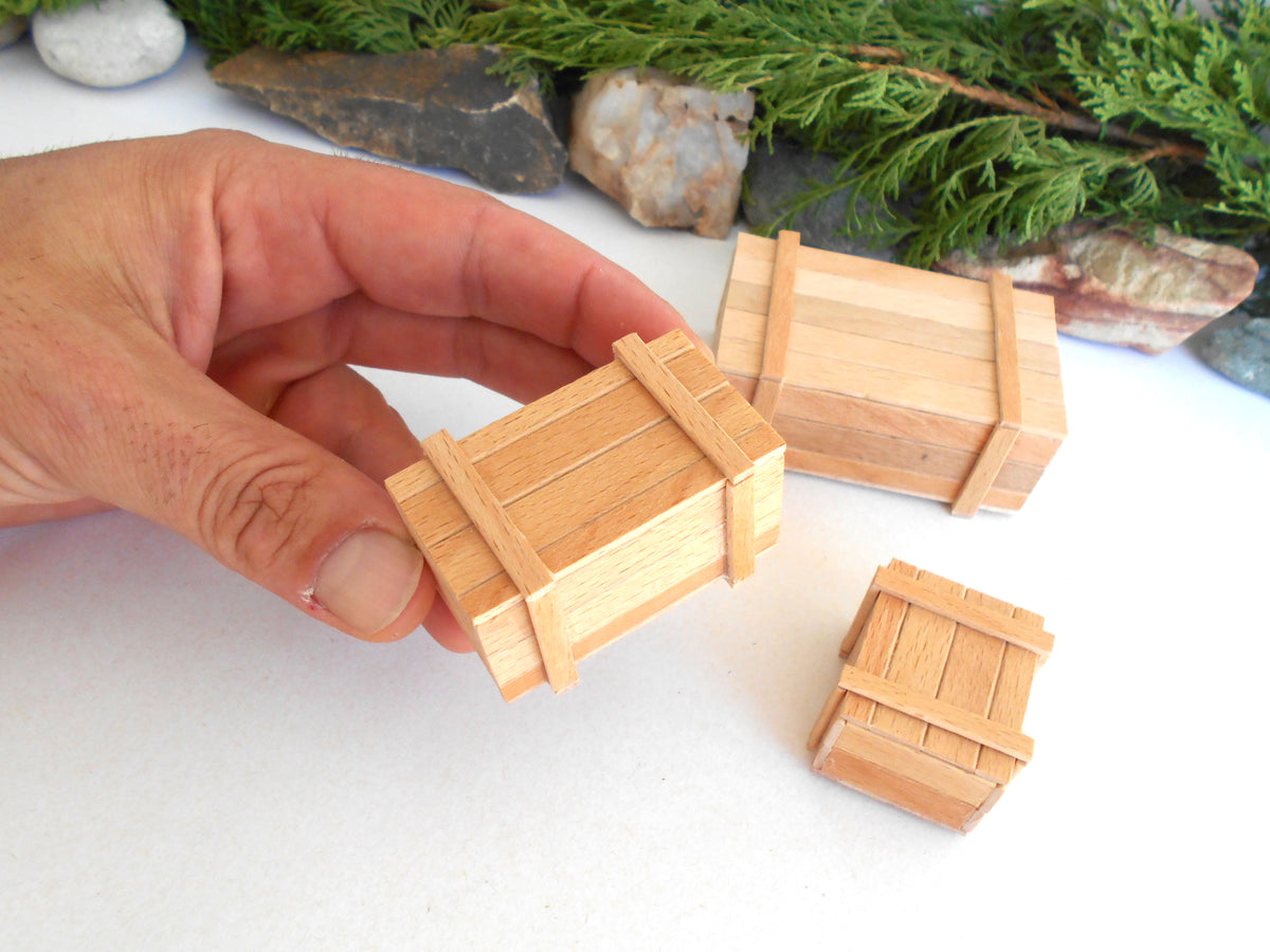 A hand holding a miniature wooden crate model, with two smaller matching crate models placed on a white surface and evergreen branches and rocks in the background.