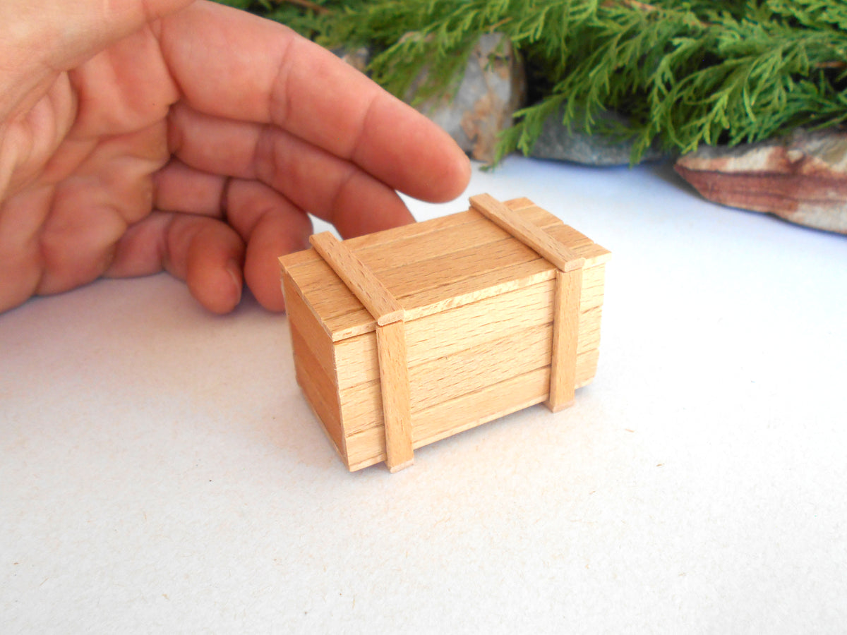 A miniature light wood crate model, shown next to a human hand to demonstrate its small scale, set against a white surface with blurred green foliage in the background.